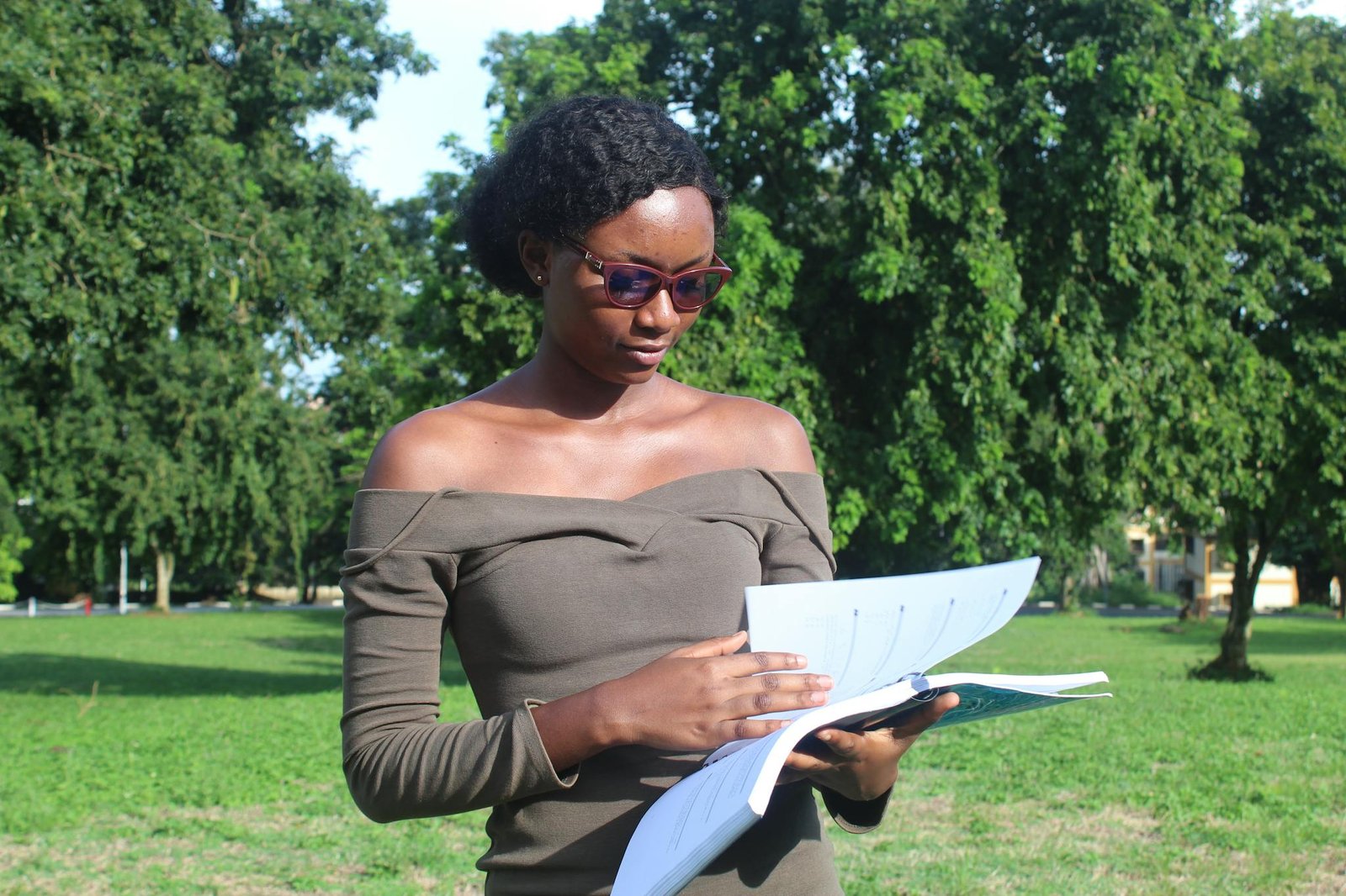 A woman reading documents in a park in Kumasi, surrounded by lush trees on a sunny day.